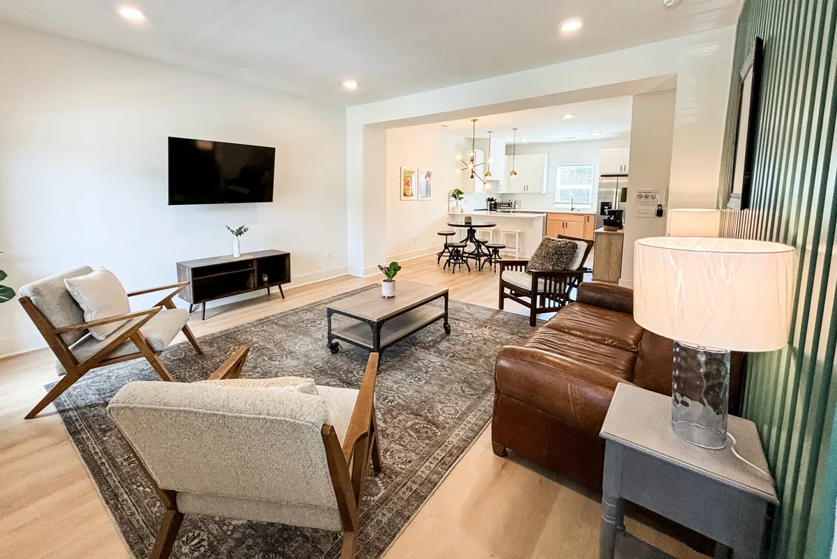 A spacious living room with a cozy sofa, multiple armchairs, a coffee table, and a large television mounted on a green accent wall. The room is styled for comfort and socializing, with tasteful decor and plenty of seating for guests.