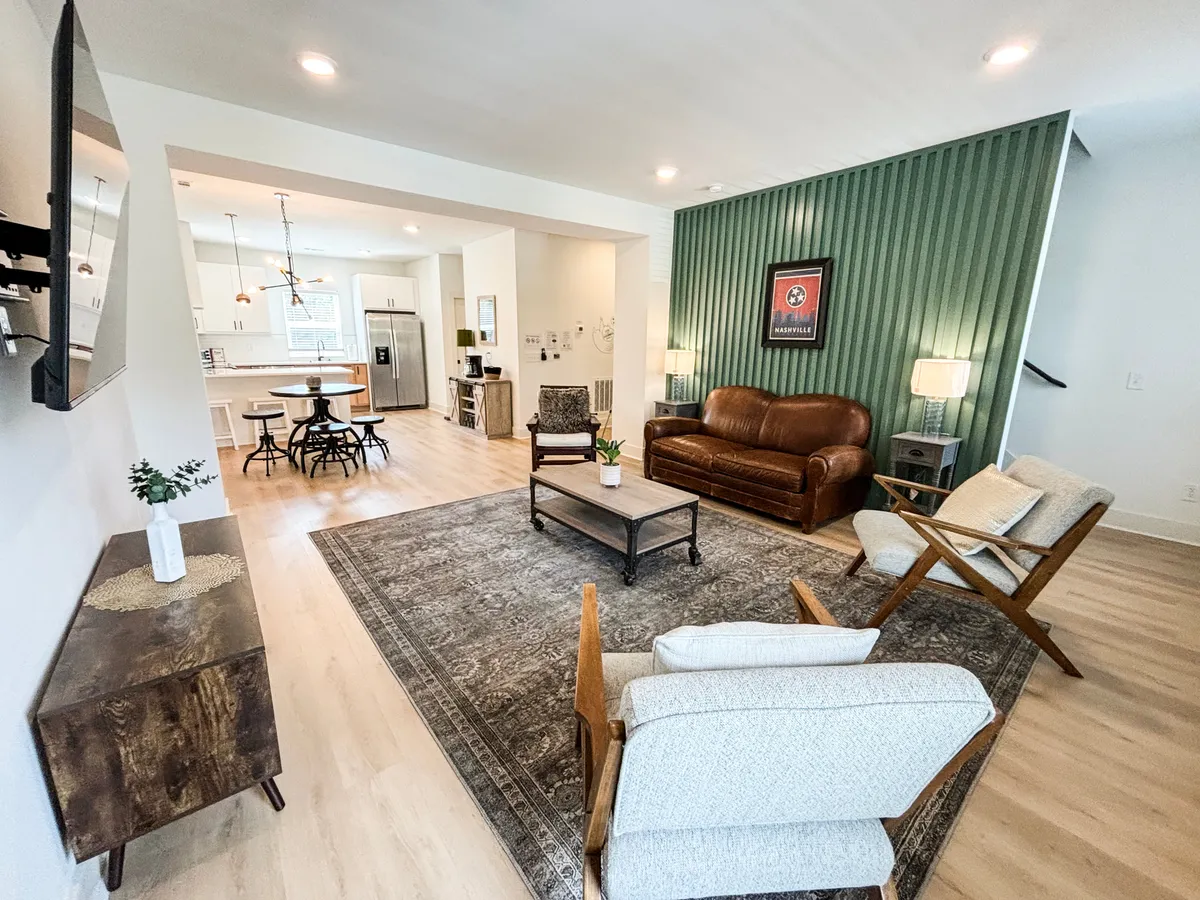 The hero image showcases a stylish, cozy living area featuring a plush leather sofa, a textured accent wall, a contemporary coffee table atop a chic area rug, and a welcoming dining area in the background. The room is arranged for both relaxation and gathering, ideal for movie nights, game time, or shared meals with family and friends.