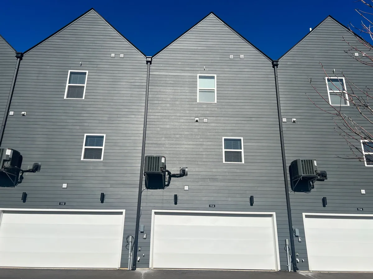 A contemporary exterior shot of a modern residential building, featuring gray siding, white garage doors, and small windows. Air conditioning units are visible, indicating comfort and practicality. The structure is part of a larger community, suggesting multiple units available.