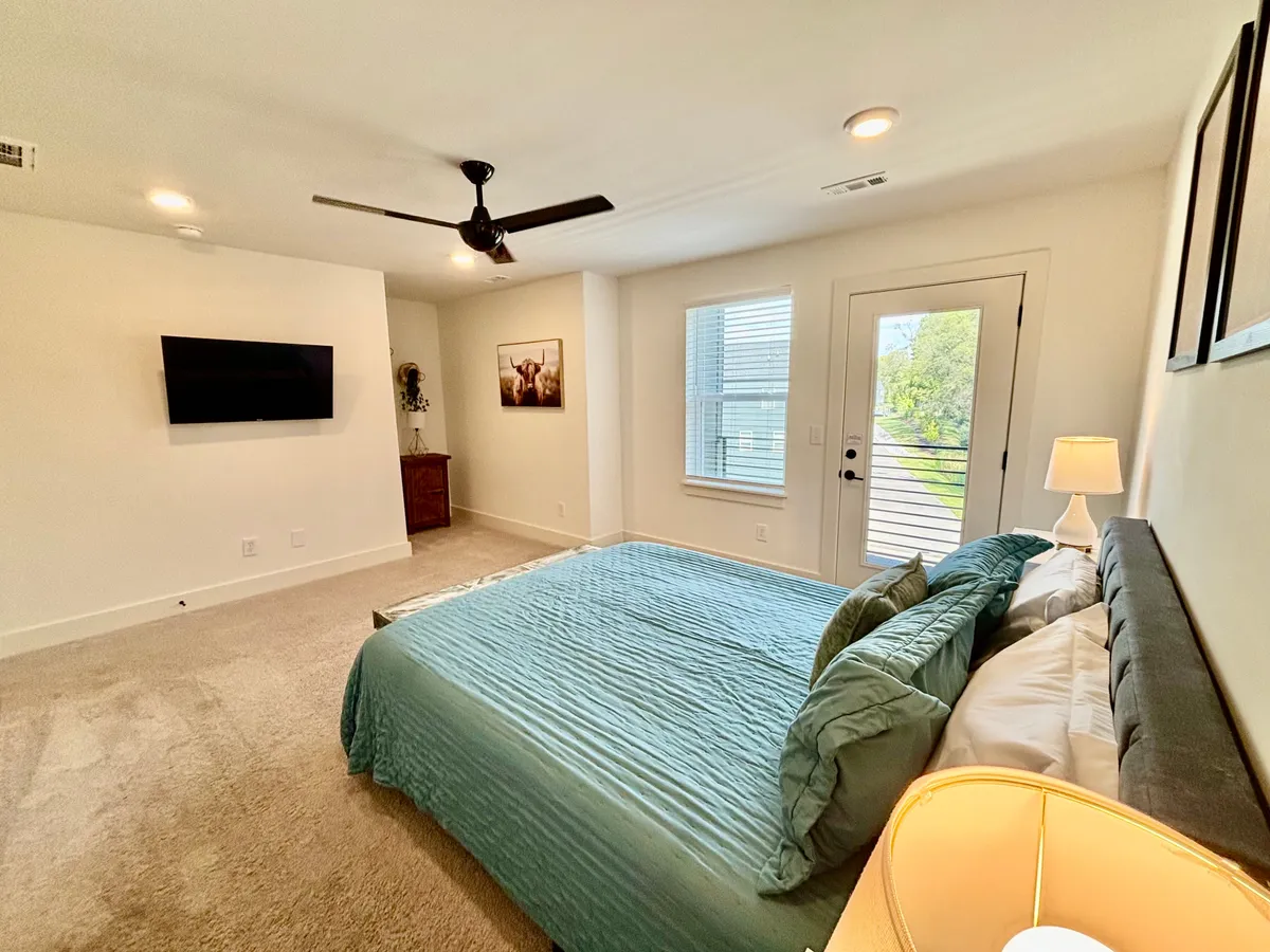 A queen bedroom featuring a bed with soft bedding, a nightstand with lamp, a television mounted on the wall, a window with natural light streaming in, and a door leading outside. The space exudes comfort and tranquility, ideal for a restful retreat.