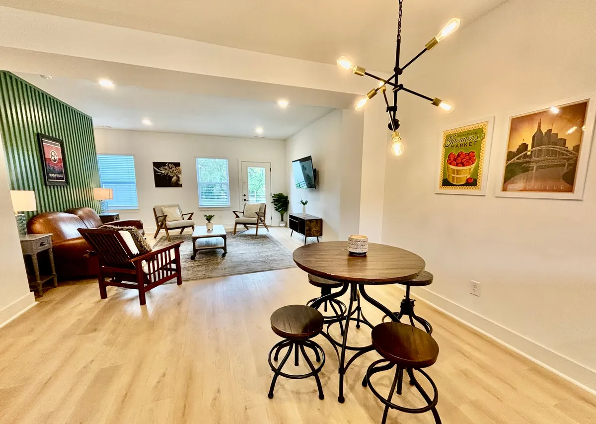 A round dining table surrounded by stools sits beneath a unique light fixture, complemented by a green accent wall and adjacent comfortable seating. The setup invites intimate dining and relaxed group conversations.