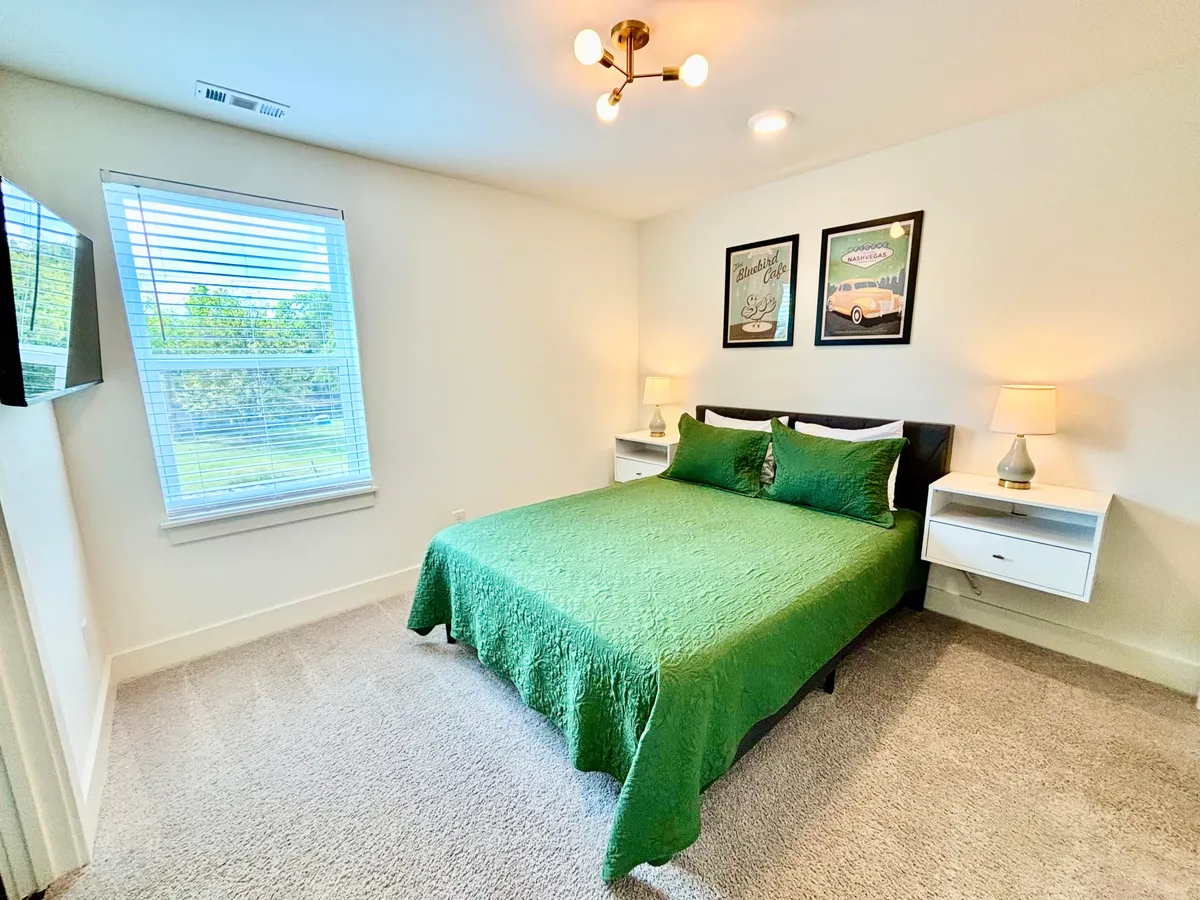 A calm and comfortable bedroom with green bedding, art prints on the wall, nightstands with lamps, and a window with blinds. The space is filled with gentle light, making it an inviting sanctuary to unwind.